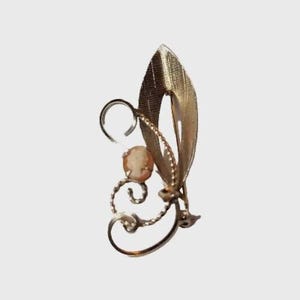 May include: A gold-toned vintage brooch with a leaf design and a cameo. The brooch features a textured leaf shape, a small cameo, and decorative swirls. The pin is a classic piece of jewelry.