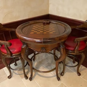 May include: A round, dark wood game table with a built-in chessboard, flanked by two matching chairs. The chairs have red leather seats and curved wooden frames. The table and chairs are in a corner setting.