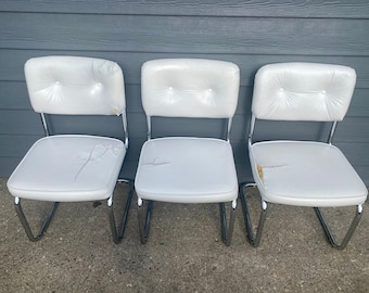 May include: Three white chairs with chrome frames. The chairs have tufted upholstery and are arranged in a row.