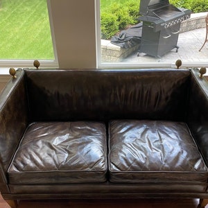May include: A dark brown leather sofa with two cushions and brass accents on the arms. The sofa is in a room with a window behind it.
