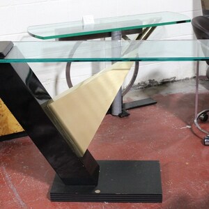 May include: A modern, geometric console table with a clear glass top. The table features a black, V-shaped support with a gold-colored accent and a rectangular base. The table's design is sleek and contemporary.