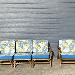 Könnte beinhalten: Vier Bambus-Stühle mit blauen Kissen und floralem Stoffmuster. Die Stühle sind in einer Reihe vor einer grauen Holzwand angeordnet.