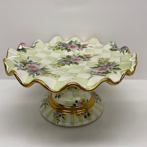May include: A white ceramic pedestal cake stand with a green and white checkered pattern and floral accents. The stand has a scalloped edge and gold trim.