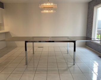 May include: A chrome and glass dining table with a rectangular top and two leaves that extend the length of the table. The table is in a room with white walls and tiled floors.