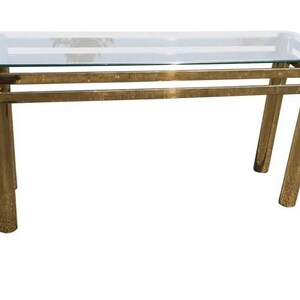 May include: A gold metal console table with a glass top. The table has a simple, modern design with a rectangular top and four legs.