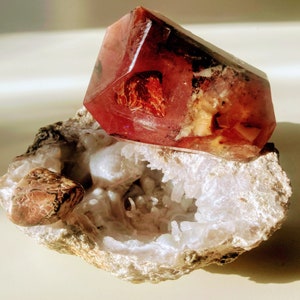 May include: A large, pink, translucent crystal with red and orange inclusions sits on top of a white, geode-like rock. The crystal is shaped like a pyramid with a flat base and a pointed top.
