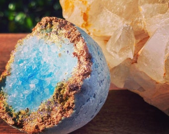 May include: A spherical bath bomb with a light blue exterior and a textured, crystalline blue center. The edge of the opening is rimmed with metallic copper and gold accents, featuring a rough, geode-like appearance with visible glitter particles.