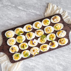 May include: A wooden serving board with a floral design engraved on the surface. The board is filled with deviled eggs, each topped with a different garnish, such as dill, parsley, and capers.