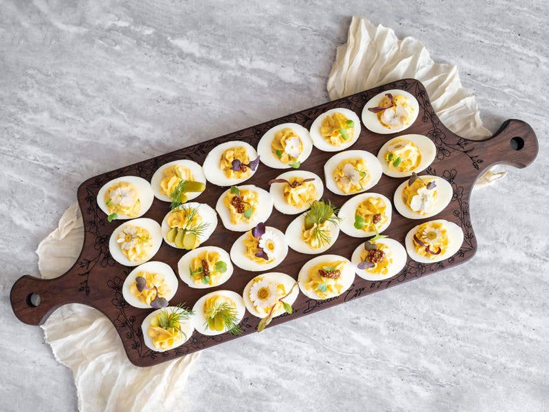 May include: A wooden serving board with a floral design engraved on the surface. The board is filled with deviled eggs, each topped with a different garnish, such as dill, parsley, and capers.