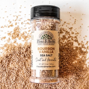 May include: A glass jar filled with brown sea salt with a brown label that reads "Flour & Herbs" and "Bourbon Vanilla Sea Salt" with the tagline "Sweet and Aromatic". The label also includes the net weight of 5 oz / 141 g and the website FlourAndHerbs.com.