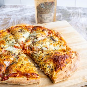 May include: A close-up of a pizza with a slice pulled away, revealing melted cheese. The pizza is topped with tomato sauce, cheese, and herbs. A small bag of pizza seasoning is visible in the background.