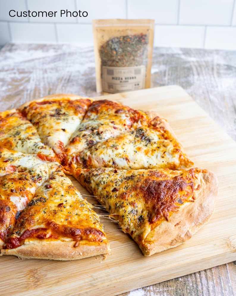 May include: A close-up of a pizza with a slice pulled away, revealing melted cheese. The pizza is topped with tomato sauce, cheese, and herbs. A small bag of pizza seasoning is visible in the background.