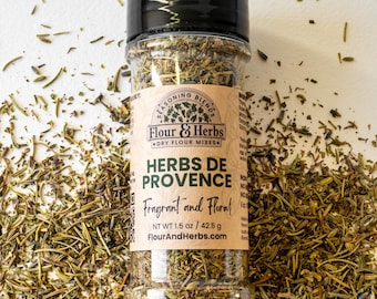 May include: A clear glass jar filled with dried herbs, labeled "Herbs de Provence Fragrant and Floral" with the brand name "Flour & Herbs Dry Flour Mixes". The jar is sitting on a white surface with scattered dried herbs.