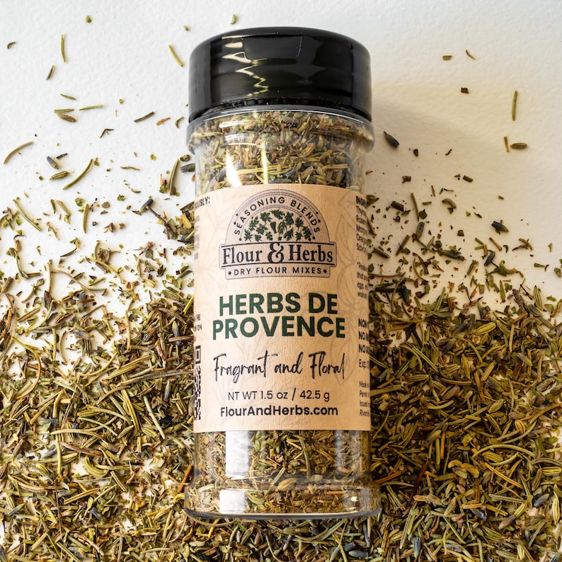 Pu&ograve; includere: Un barattolo di vetro trasparente pieno di erbe secche, etichettato "Herbs de Provence Fragrant and Floral" con il marchio "Flour & Herbs Dry Flour Mixes". Il barattolo &egrave; su una superficie bianca con erbe secche sparse.