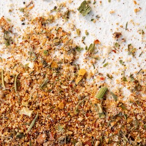 May include: Close-up of a blend of dried herbs and spices, including green herbs, red pepper flakes, and yellow flakes. The mixture is on a white surface.