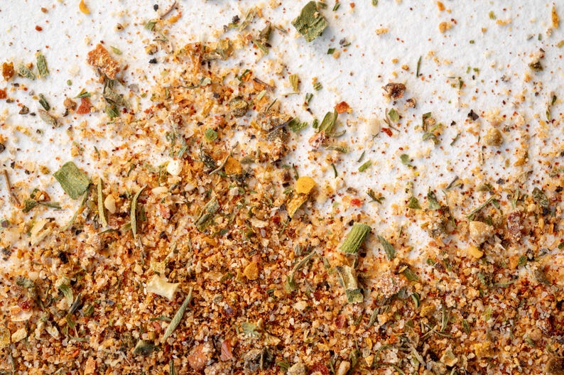 May include: Close-up of a blend of dried herbs and spices, including green herbs, red pepper flakes, and yellow flakes. The mixture is on a white surface.