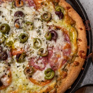 May include: A close-up of a pizza with a golden brown crust, topped with mozzarella cheese, pepperoni, green olives, and a green sauce. The pizza is in a cast iron skillet.