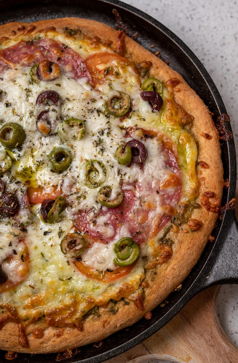 May include: A close-up of a pizza with a golden brown crust, topped with mozzarella cheese, pepperoni, green olives, and a green sauce. The pizza is in a cast iron skillet.