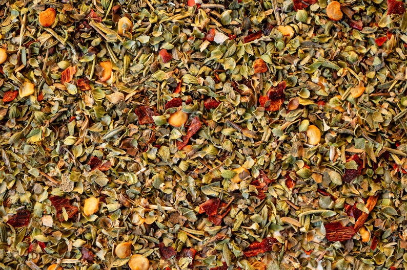 May include: A close-up of a blend of dried herbs and spices, including green herbs, red pepper flakes, and small yellow seeds.