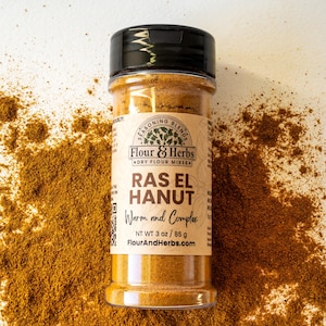 May include: A glass jar of Ras El Hanut spice blend, labeled "Flour & Herbs Dry Flour Mixes" with the text "Warm and Complex" and "NT WT 3 oz / 85 g" on the label. The jar is sitting on a white surface with brown spice scattered around it.