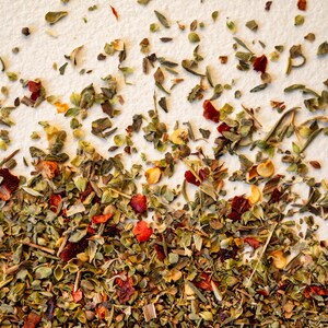 May include: A close-up of a mixture of dried herbs and spices, including green herbs, red pepper flakes, and yellow seeds. The mixture is spread out on a white surface.