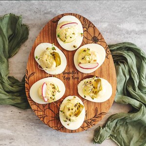 May include: A wooden egg tray with a floral design and the text "Danielle's Egg Tray" engraved on it. Six deviled eggs are arranged in the tray, topped with dill, radish slices, and a pickle spear.