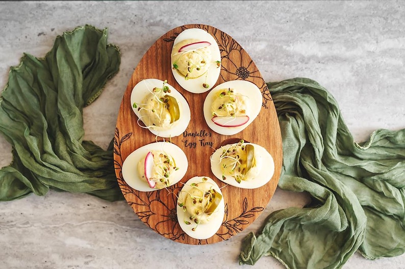 May include: A wooden egg tray with a floral design and the text "Danielle's Egg Tray" engraved on it. Six deviled eggs are arranged in the tray, topped with dill, radish slices, and a pickle spear.