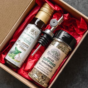 May include: A gift set in a cardboard box with red tissue paper includes a bottle of olive oil, a bottle of bread dipping seasoning, and a stainless steel pour spout. The olive oil bottle is labeled "California Olive Oil Mediterranean Medley".