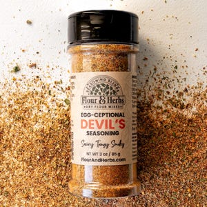 May include: A glass jar of brown seasoning blend labelled "Egg-Ceptional Devil's Seasoning Savory Tangy Smoky" with the brand name "Flour & Herbs" and the website "FlourAndHerbs.com". The jar is on a white surface with a scattering of the seasoning blend around it.