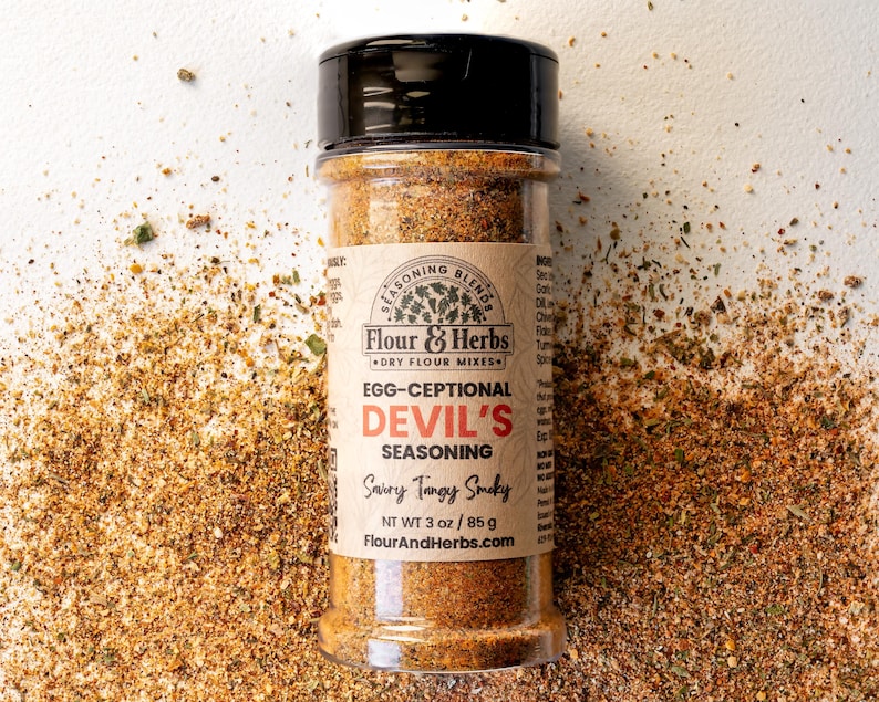 May include: A glass jar of brown seasoning blend labeled "Egg-Ceptional Devil's Seasoning Savory Tangy Smoky" with the brand name "Flour & Herbs" and the website "FlourAndHerbs.com". The jar is on a white surface with a scattering of the seasoning blend around it.