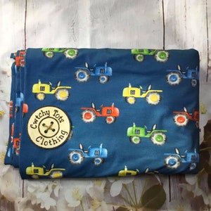 Può includere: Tessuto blu con un motivo ripetuto di trattori giocattolo gialli, rossi, verdi e blu. Il tessuto ha un bottone con il testo "Cwtchy Tots Clothing" su di esso.