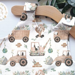 May include: A light-colored romper with a construction vehicle pattern. The romper features illustrations of dump trucks, excavators, and bulldozers in shades of brown and beige, with accents of green and white. The romper is hanging on a wooden hanger.