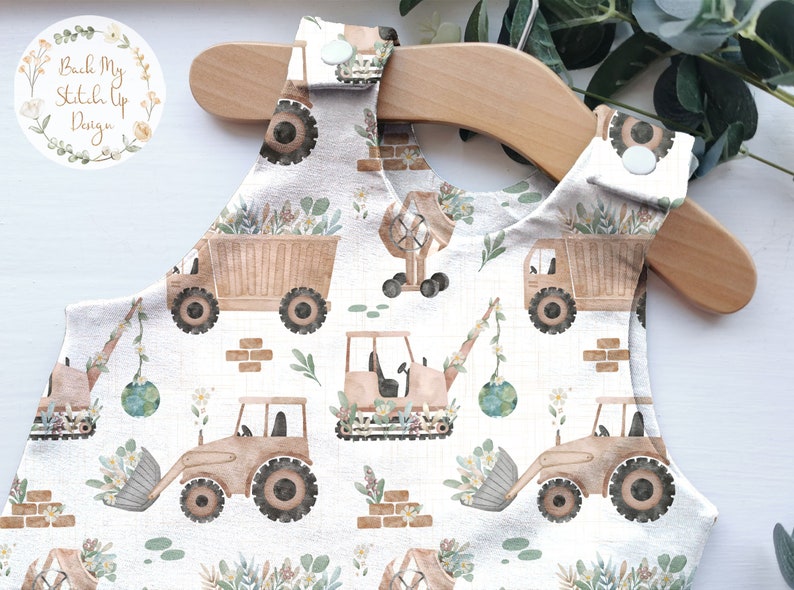 May include: A light-colored romper with a construction vehicle pattern. The romper features illustrations of dump trucks, excavators, and bulldozers in shades of brown and beige, with accents of green and white. The romper is hanging on a wooden hanger.