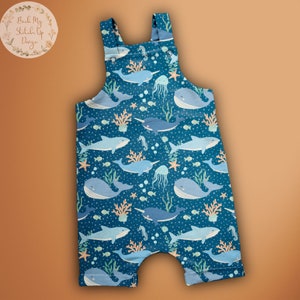 Peut inclure: Salopette bleue avec un imprimé marin fantaisiste représentant des baleines, des narvals, des méduses, des hippocampes et des coraux. Le tissu a un motif de pois blancs.