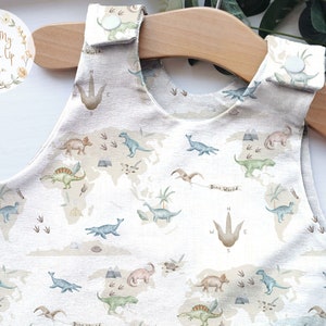 K&ouml;nnte beinhalten: Ein beiger Baby-Strampler mit einem Dinosaurier-Weltkarten-Design. Der &auml;rmellose Strampler zeigt verschiedene Dinosaurier, Fu&szlig;abdr&uuml;cke und den Text "Dino World". Der Stoff ist hellfarbig mit einem strukturierten Aussehen.