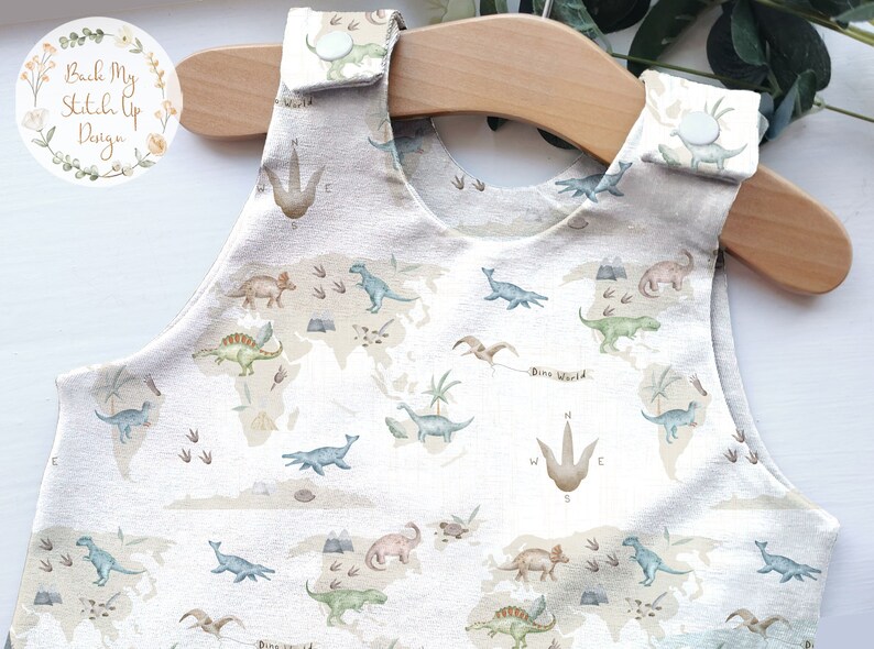 K&ouml;nnte beinhalten: Ein beiger Baby-Strampler mit einem Dinosaurier-Weltkarten-Design. Der &auml;rmellose Strampler zeigt verschiedene Dinosaurier, Fu&szlig;abdr&uuml;cke und den Text "Dino World". Der Stoff ist hellfarbig mit einem strukturierten Aussehen.