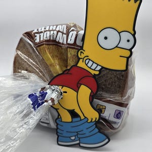 May include: A novelty magnet featuring a cartoon character with yellow skin, a red shirt, and blue shoes. The magnet is placed on a loaf of whole wheat bread in a plastic bag. The character is in a humorous pose.