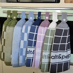 May include: A row of eight hanging kitchen towels with crocheted tops and button fastenings. The towels feature various patterns including checkered and grid designs in shades of green, blue, navy, lavender, white, and grey, with text reading alt&pep.