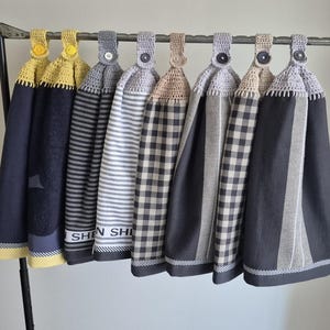 May include: A selection of hanging kitchen towels in various patterns and colours. The towels are displayed on a metal rack. Designs include stripes, gingham, and solid colours, with crocheted tops and button details. The colour palette includes navy, grey, and yellow.