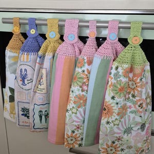 May include: A selection of seven hanging kitchen towels with crochet tops and decorative buttons. The towels display diverse patterns, including floral, striped, and illustrated designs. The crochet tops are in shades of yellow, blue, pink, and green.