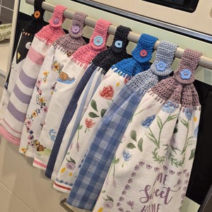 May include: A row of eight hanging kitchen towels with crocheted tops in various colours, each secured with a button. The towels feature diverse patterns including floral prints, plaid, and a graphic with the text Home Sweet Home, all suspended from a horizontal bar.