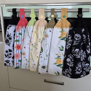 May include: Seven decorative kitchen towels hanging on a metal bar. Each towel has a unique floral design, with crochet tops in various colours such as black, red, green, and yellow. The towels are made of fabric with different floral patterns.
