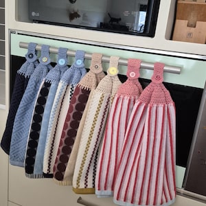 May include: A row of eight hanging kitchen towels with crocheted tops, each secured by a single button. The towels feature various patterns including geometric shapes, polka dots, and vertical stripes in blue, cream, pink, and red colour palettes.
