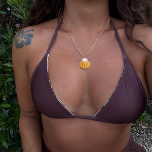 May include: A gold-toned chain necklace featuring a small, fan-shaped yellow and white seashell pendant. The necklace is worn with a dark brown bikini top that has thin halter-style straps and a simple triangular design.