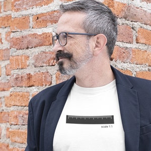 Op de afbeelding: Een wit T-shirt met een zwarte liniaal afbeelding en de tekst "schaal 1:1" gedrukt op de voorkant. De persoon die het shirt draagt staat voor een bakstenen muur.