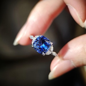 Può includere: Primo piano di un anello d'argento con una grande pietra preziosa blu scuro a taglio quadrato. L'anello è adornato con pietre preziose più piccole e trasparenti attorno alla pietra principale e lungo la fascia, creando un effetto scintillante.