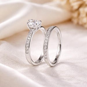 May include: A pair of silver rings, one with a large, round, clear gemstone and the other with a band of smaller, clear gemstones. The rings are set against a soft, white background, suggesting a jewelry or wedding theme.