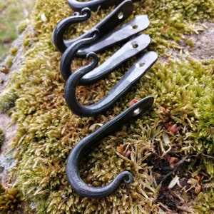 Op de afbeelding: Een set van vijf handgemaakte, zwarte metalen haken met een gehamerde textuur. Elke haak heeft een gebogen, opgerold uiteinde en een platte, rechthoekige basis met een enkel gat voor montage. De haken worden getoond op een bed van groen en bruin mos.