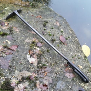 May include: A long, thin, black metal tool with a curved handle and a flat, pointed end. The tool is lying on a rock covered in moss and leaves.
