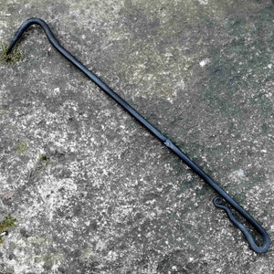 Könnte beinhalten: Ein schwarzer Metall-Kaminbesen mit einem gebogenen Griff. Der Besen liegt auf einer grauen, strukturierten Oberfläche.